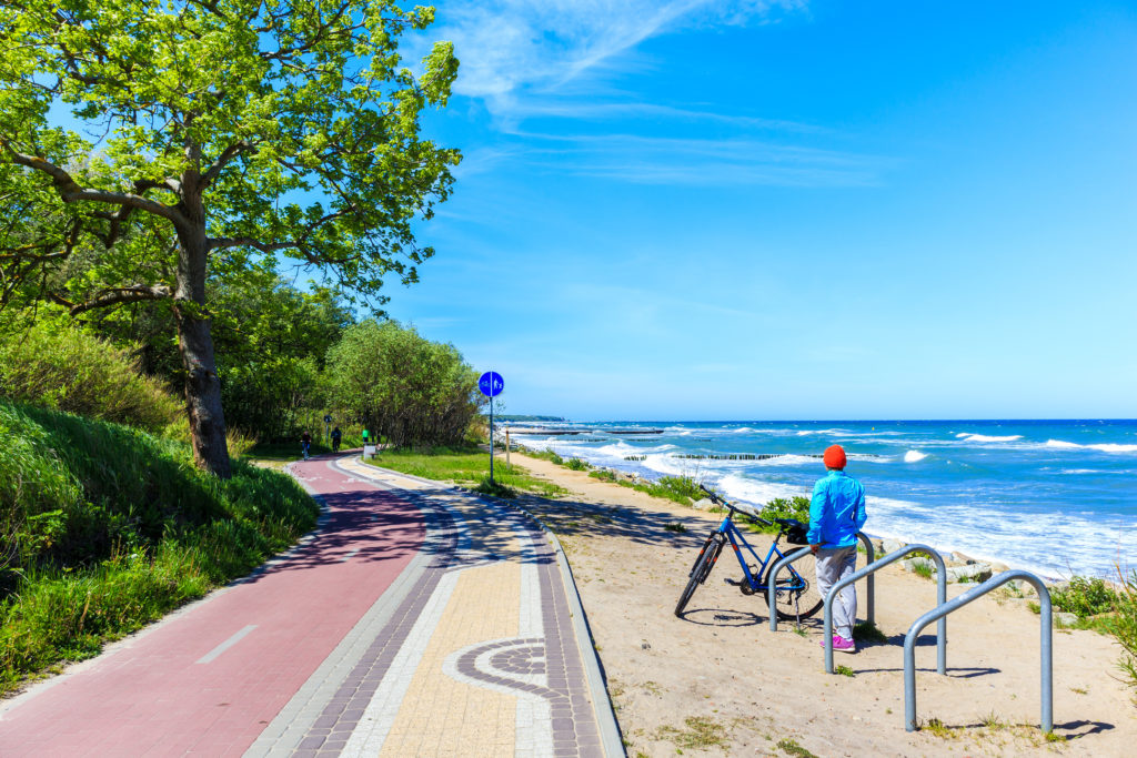 Kobieta z rowerem na plaży obok ścieżki rowerowej patrząca na morze. 